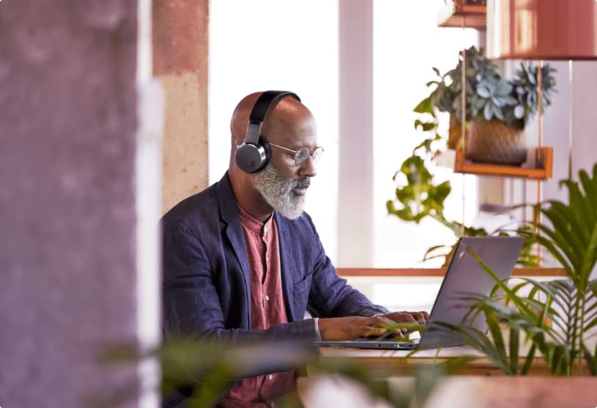 A bearded man in headphones types on a laptop at a desk surrounded by plants.