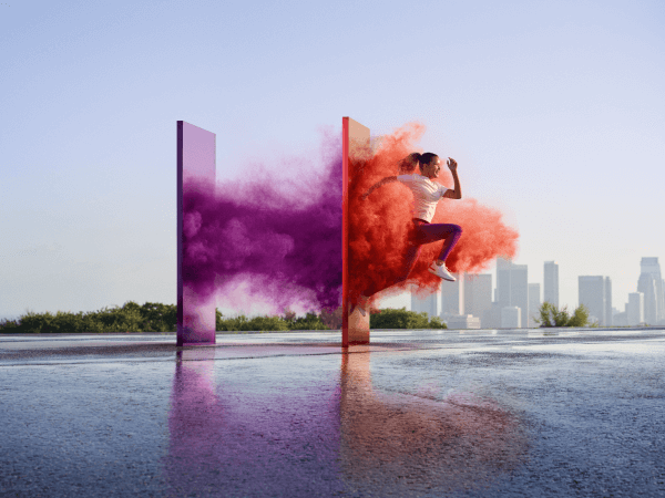 Un monolito morado y un monolito naranja se elevan sobre un cuerpo de agua con el horizonte de una ciudad de fondo mientras una mujer parece correr sobre el agua, atravesando los monolitos y creando humo morado y naranja.