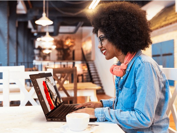 A woman smiles while sitting at a table or desk in an open, lofted workspace and typing on a Lenovo ThinkPad mobile workstation with a cup of coffee next to it.
