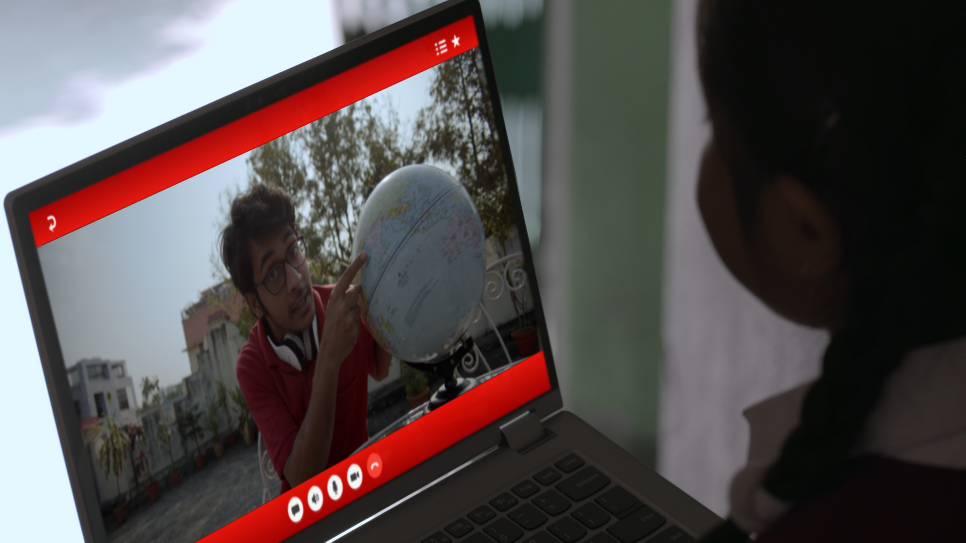 A student taking her online lessons on a Lenovo laptop, while her volunteer teacher is seen pointing to a globe
