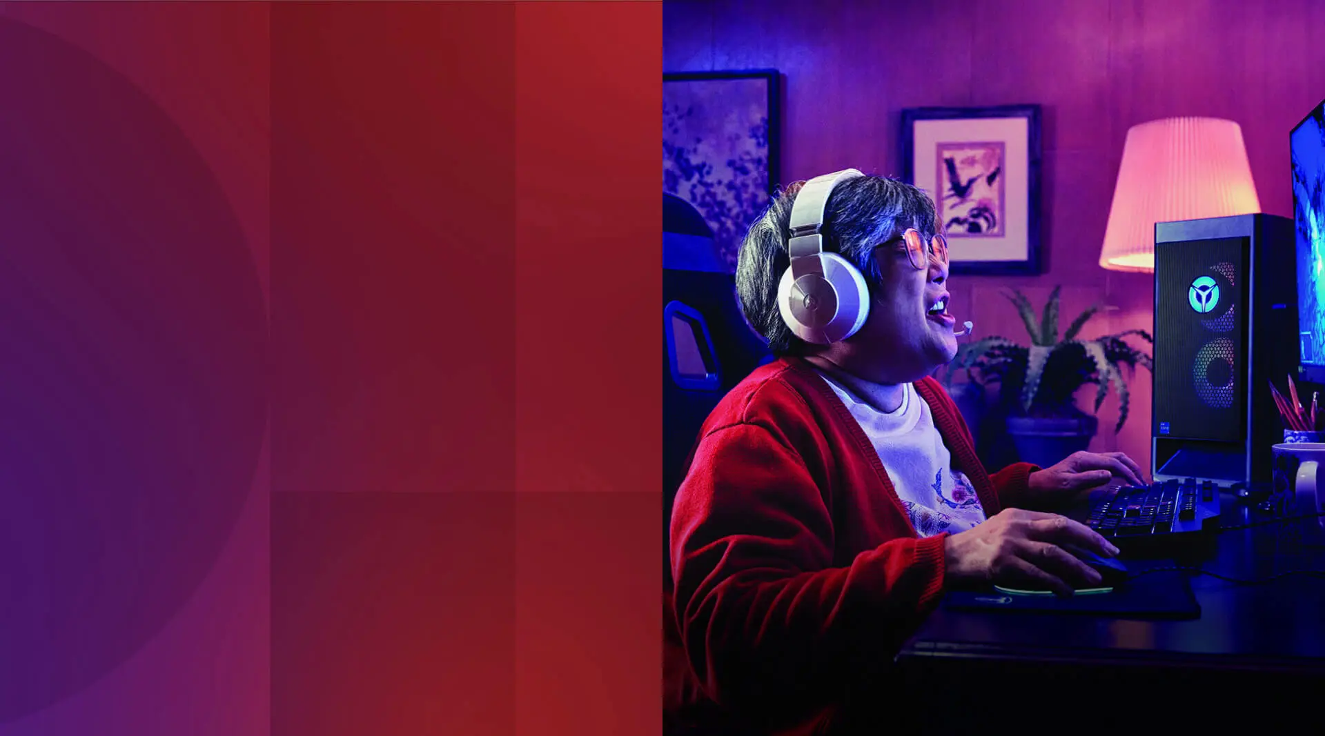 In a violet-lit room, a woman with salt-and-pepper hair, glasses, and a big gaming headset engages her keyboard and mouse as she plays a computer game. Papers, a coffee cup, and a container of writing utensils also occupy the desk.