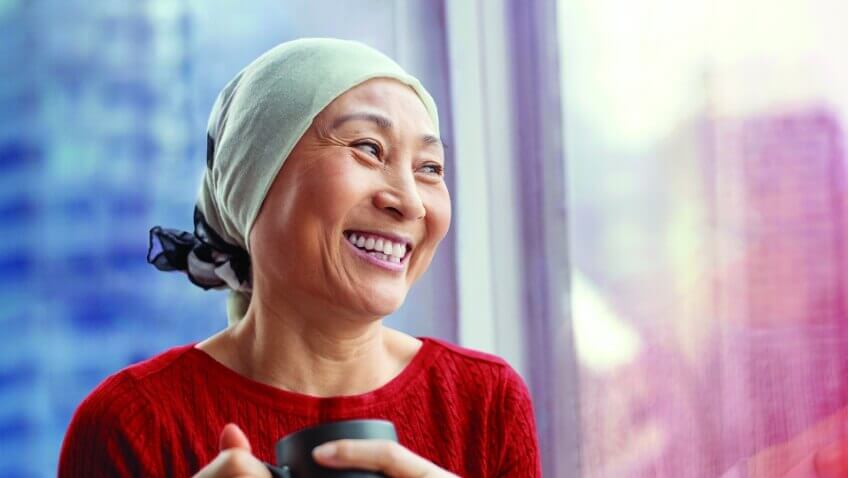 A gently smiling young girl in a red-striped shirt looks down upon a pale bedridden woman with a shaved head and an exuberant smile.