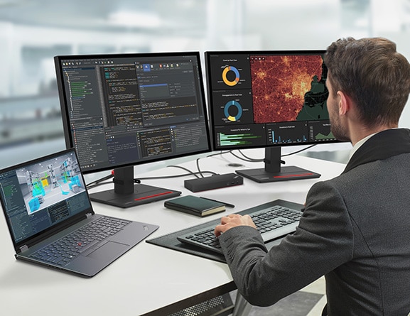 Gaming developer looking at code on two large monitors, with an opened Lenovo ThinkPad P16 Gen 2 (16″ Intel) laptop to the left