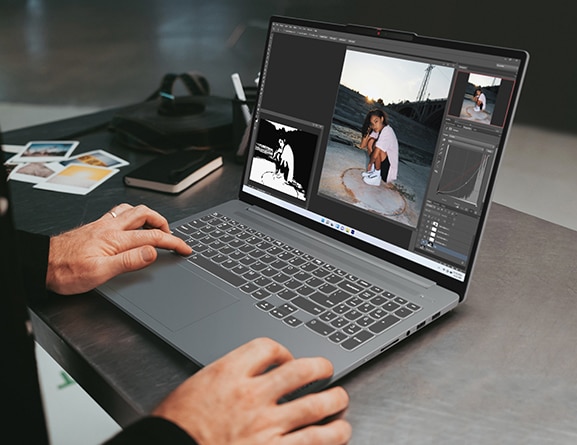 A pair of hands works on the IdeaPad Pro 5 Gen 8 (16'' intel), which rests on a small black table next to photos and notebook