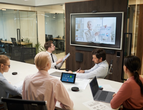 5 people sitting around a conference table with a 6th person video chatting on wall-mounted TV, with Cam above and Bar below.