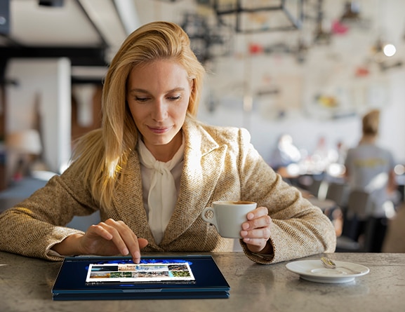 Head-on view of a woman in a coffee shop using the touchscreen of an abyss blue ThinkBook 14s Yoga Gen 2 convertible laptop that's in tablet mode on the table in front of her.
