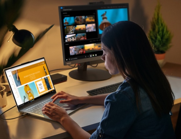 Eine Frau arbeitet bei schwachem Licht an einem Schreibtisch und tippt auf einem Lenovo ThinkBook 13x Notebook, das um 110 Grad aufgeklappt und an einen großen externen Monitor angeschlossen ist.