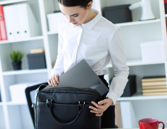 Eine Frau steht in einer hellen Büroumgebung neben einem Schreibtischstuhl und verstaut ein flaches, leichtes Lenovo ThinkBook 13x Notebook in der Farbe Storm Gray in eine schlanke Aktentasche.