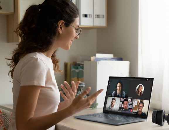 Femme souriante et faisant des gestes des mains face à l'écran d'un ordinateur portable Lenovo ThinkBook 13x posé devant elle sur un bureau, en vidéoconférence avec cinq collègues.
