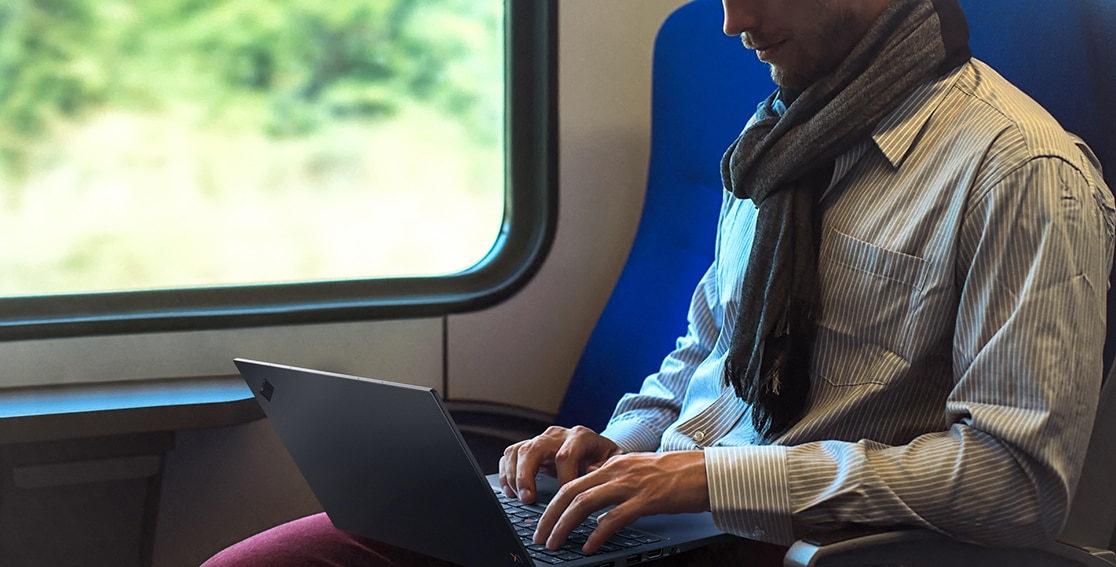 Hipster guy on train with Lenovo ThinkPad X1 Carbon in his lap in use typing on keyboard.