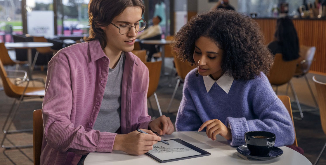 Deux étudiants dans un café avec une liseuse Lenovo Smart Paper E-Ink posée à plat, un des étudiants prenant des notes sur l’écran avec un Lenovo Smart Pen