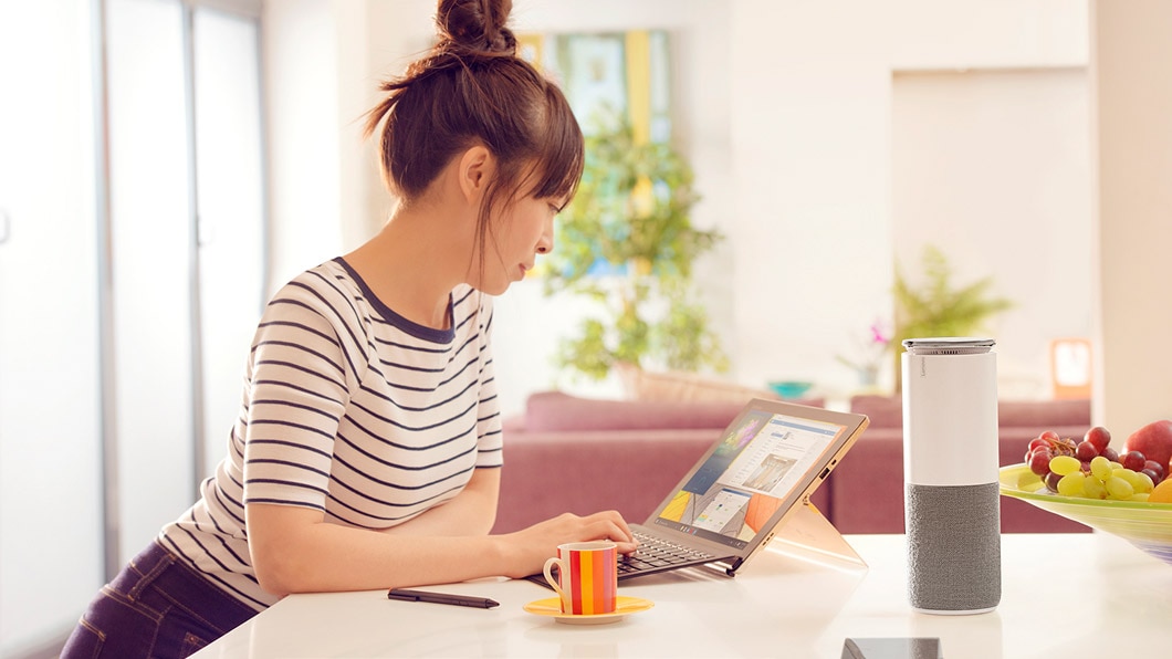 Shot of the Lenovo Smart Assistant on a kitchen surface, with a woman using a Lenovo 2-in-1 laptop