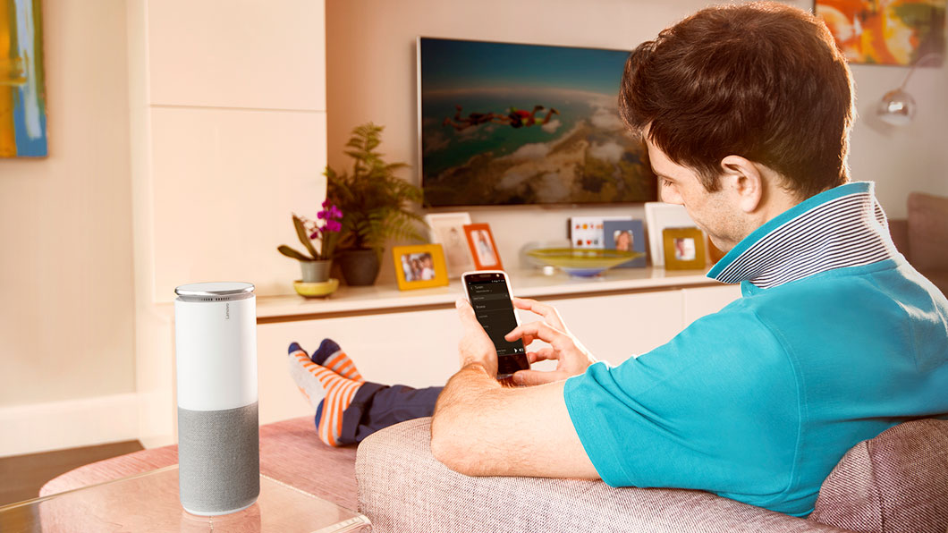 Shot of the Lenovo Smart Assistant on a side table, with a man on a sofa using a smartphone