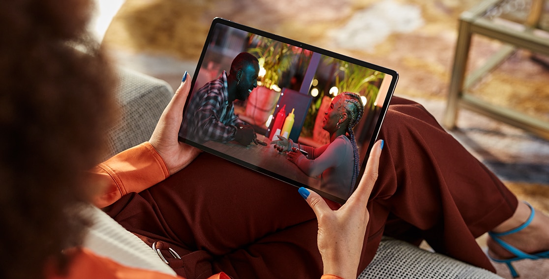 A person sits on a patio couch, facing away, while watching a colorful movie scene unfold on the Lenovo Tab P12 Pro she's holding on her lap.