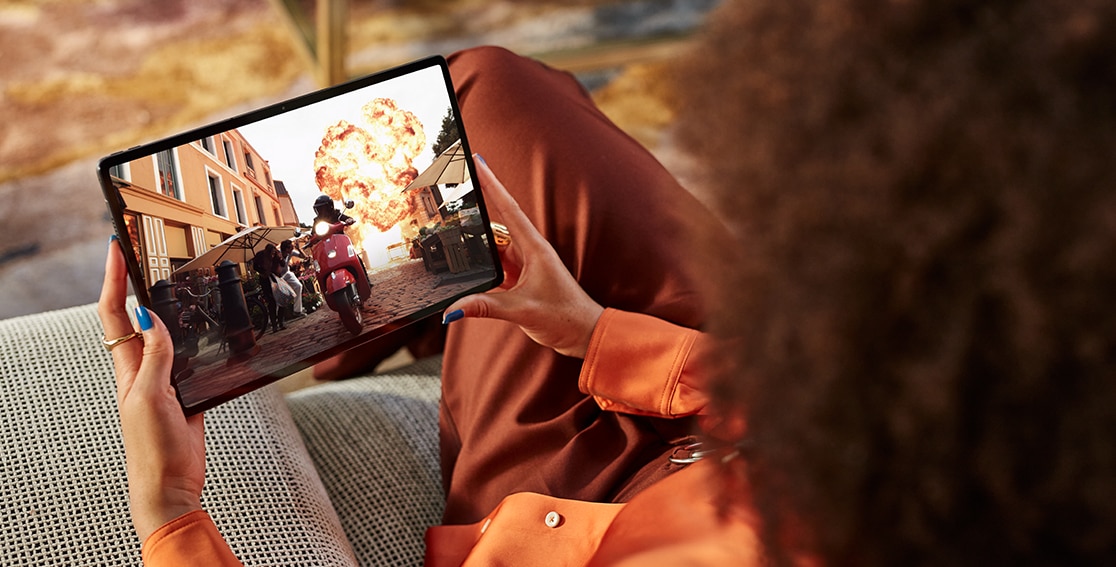 An explosive movie scene fills the Lenovo Tab P12 Pro screen as the viewer, sitting on a patio couch, holds it in her lap and enjoys the great audio.