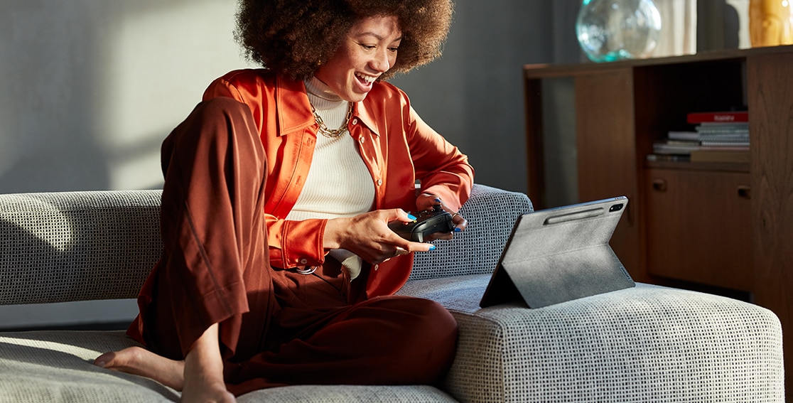 Una persona sostiene un mando de juego mientras ve cómo se desarrolla la acción en la Tablet Lenovo Tab P12 Pro que está apoyada en su soporte sobre un cojín de sofá cercano.
