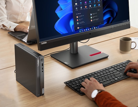 Close up of Lenovo ThinkCentre Neo 50q Gen 4 Tiny (Intel), stood vertically on an office desk, next to monitor & keyboard (sold separately), with person typing on keyboard