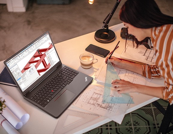 Aerial view of architect at work, with Lenovo ThinkPad P16v (16” Intel) mobile workstation showing designs in screen, and paper drawings/designs on desk