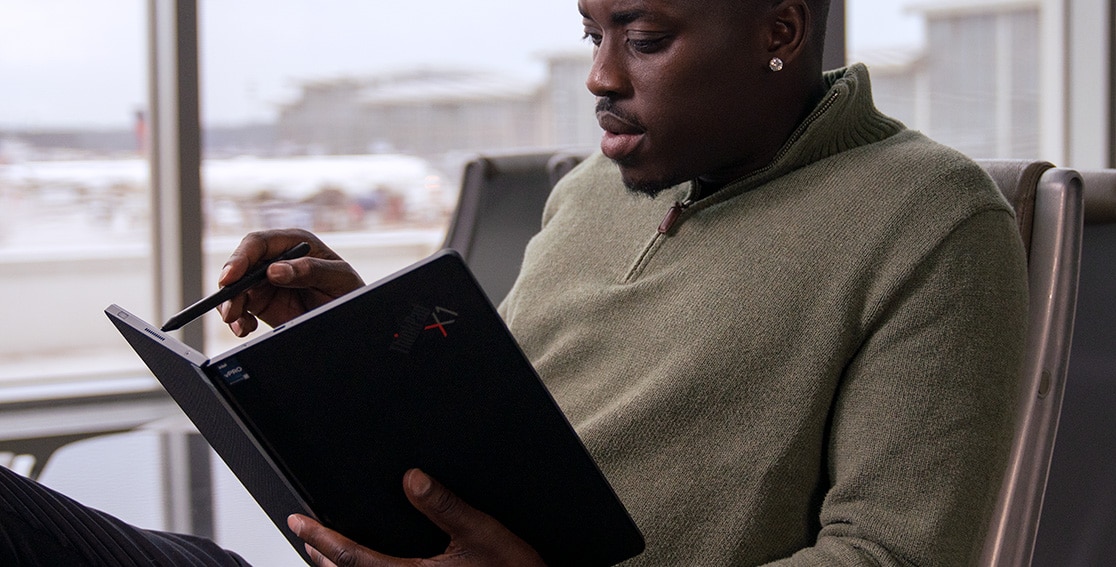 A man sits in an airport chair using the Lenovo ThinkPad X1 Fold, which he holds folded open like a book, and he’s using a pen on the right side of the display.