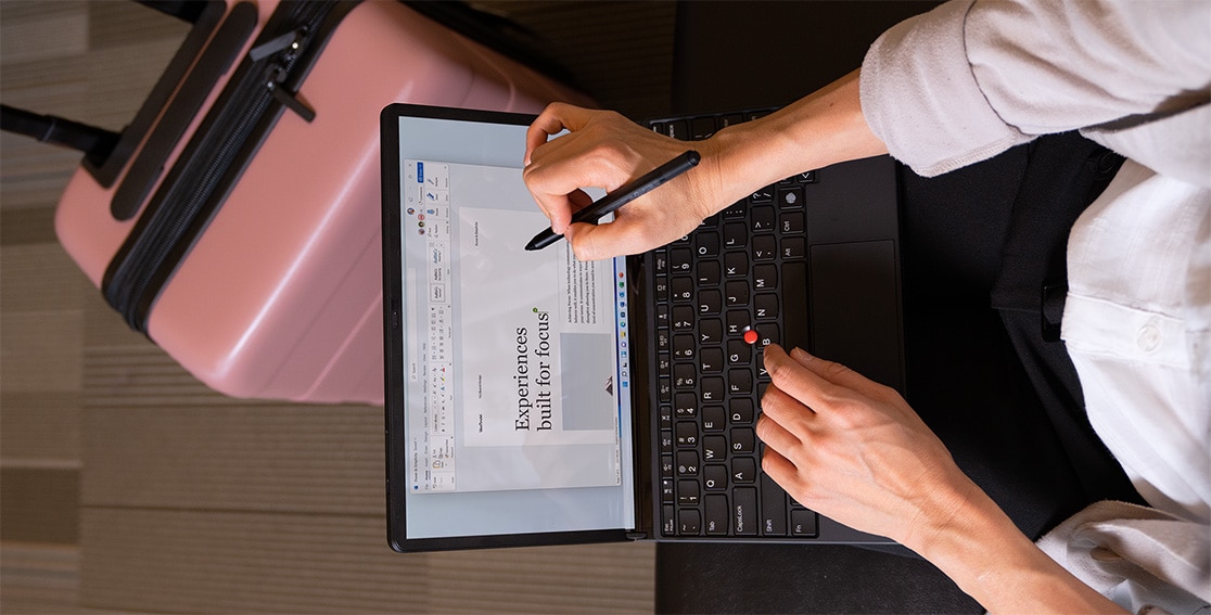 Overhead shot of a woman with the Lenovo ThinkPad X1 Fold in use on her lap with the TrackPoint Keyboard attached, as she uses a pen to make notes in the Word doc on the display.  