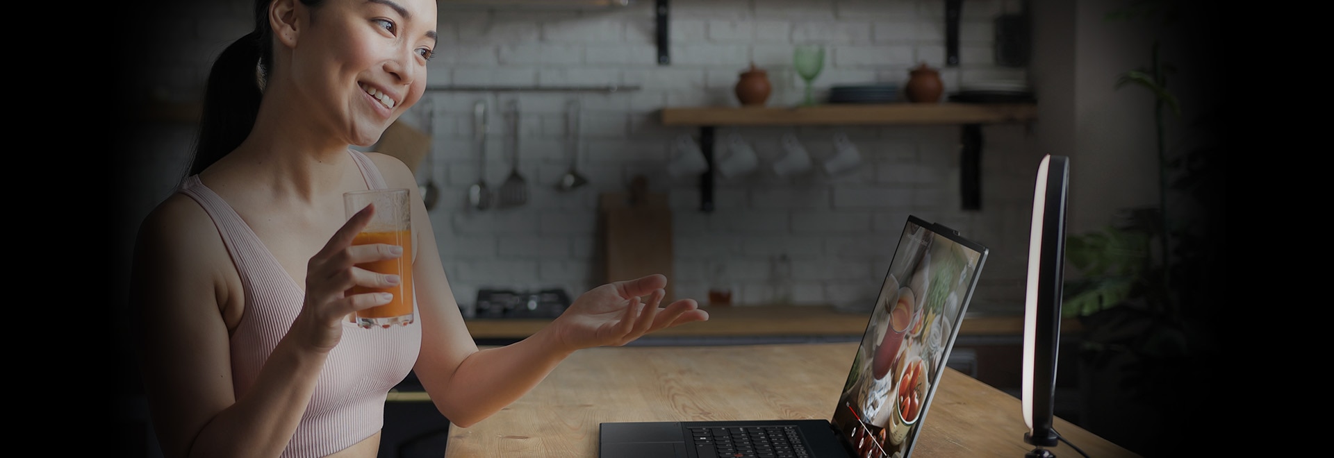Woman sitting at a table holding a glass of juice in one hand and gesturing toward the Lenovo ThinkPad Z16 laptop in front of her. 